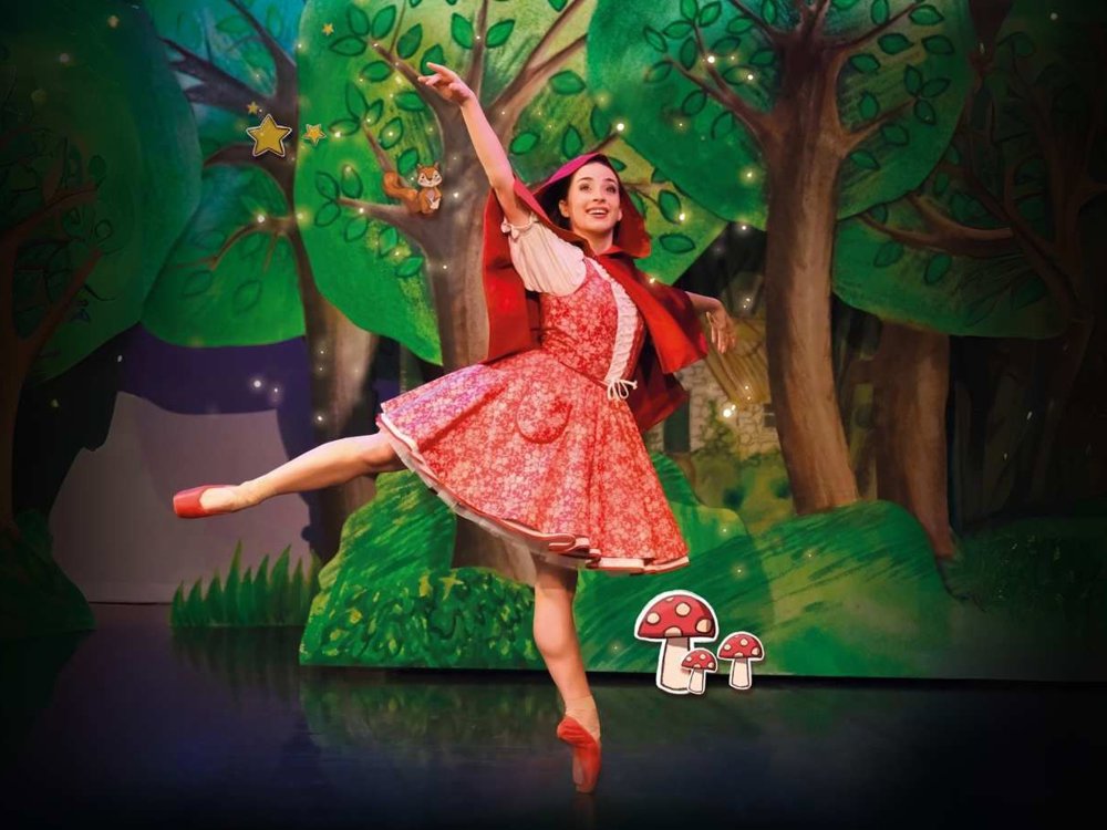 A ballet dancer dressed as Little Red Riding Hood performs a graceful pose on stage, surrounded by a colourful forest backdrop with illustrated trees, stars, and toadstools.