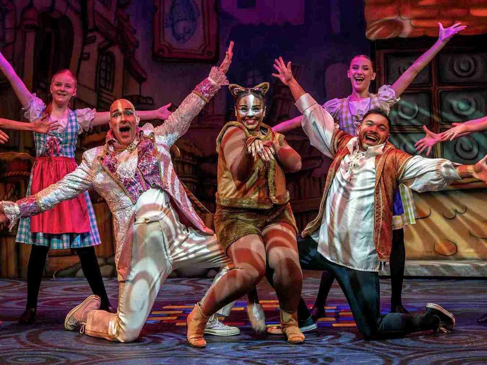 A lively stage scene featuring three central performers and supporting cast in a colourful and energetic pose. In the middle, a performer dressed as a cat strikes a playful clawing gesture, wearing a brown furry costume with cat ears. On the left, a character in a glittering white suit with pink accents beams enthusiastically with arms outstretched. On the right, a character in a medieval-inspired tunic leans forward, laughing with arms open wide. 