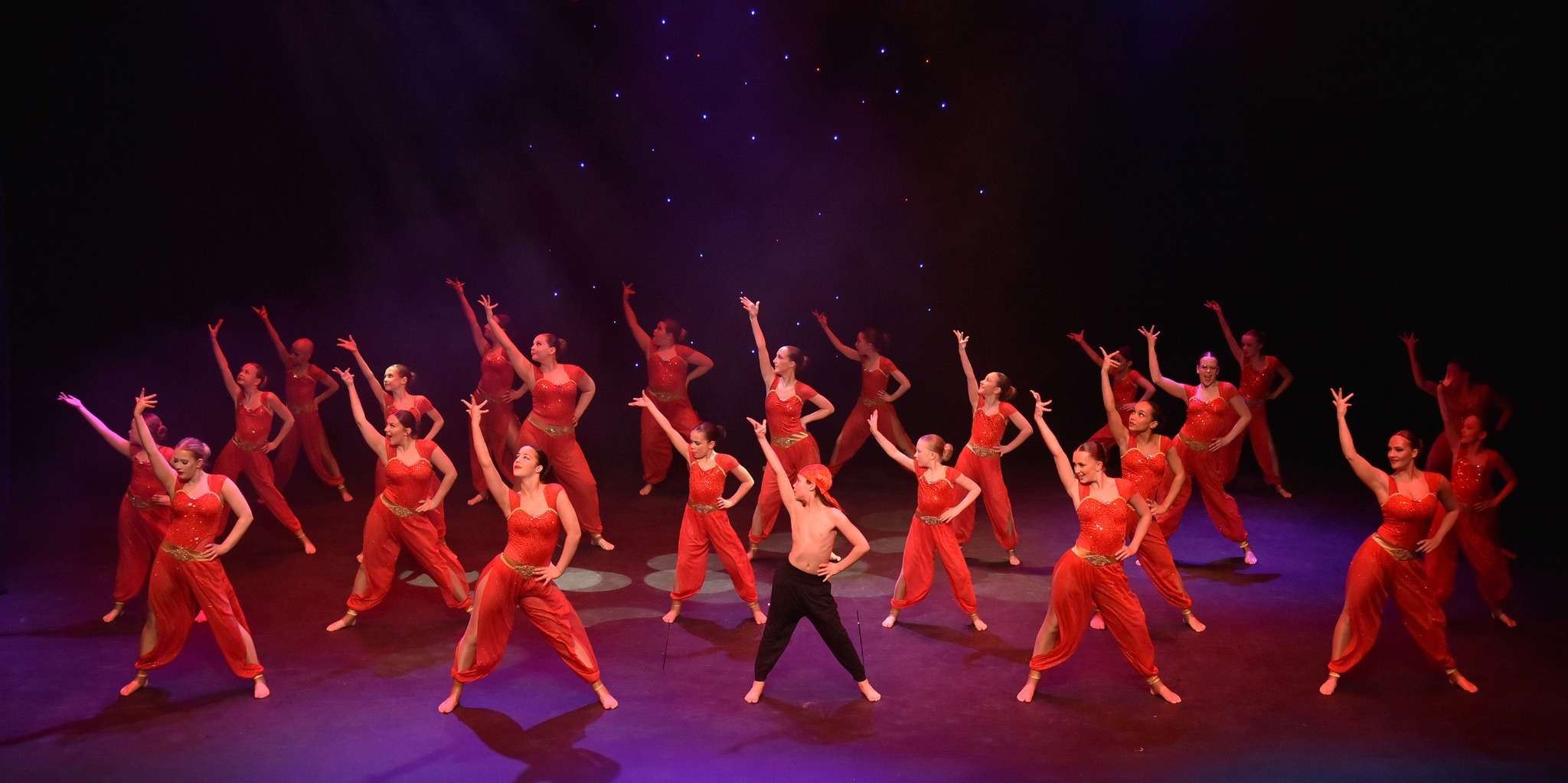 A large group of dancers performing a choreographed routine on stage, wearing red costumes and striking dramatic poses under theatrical lighting