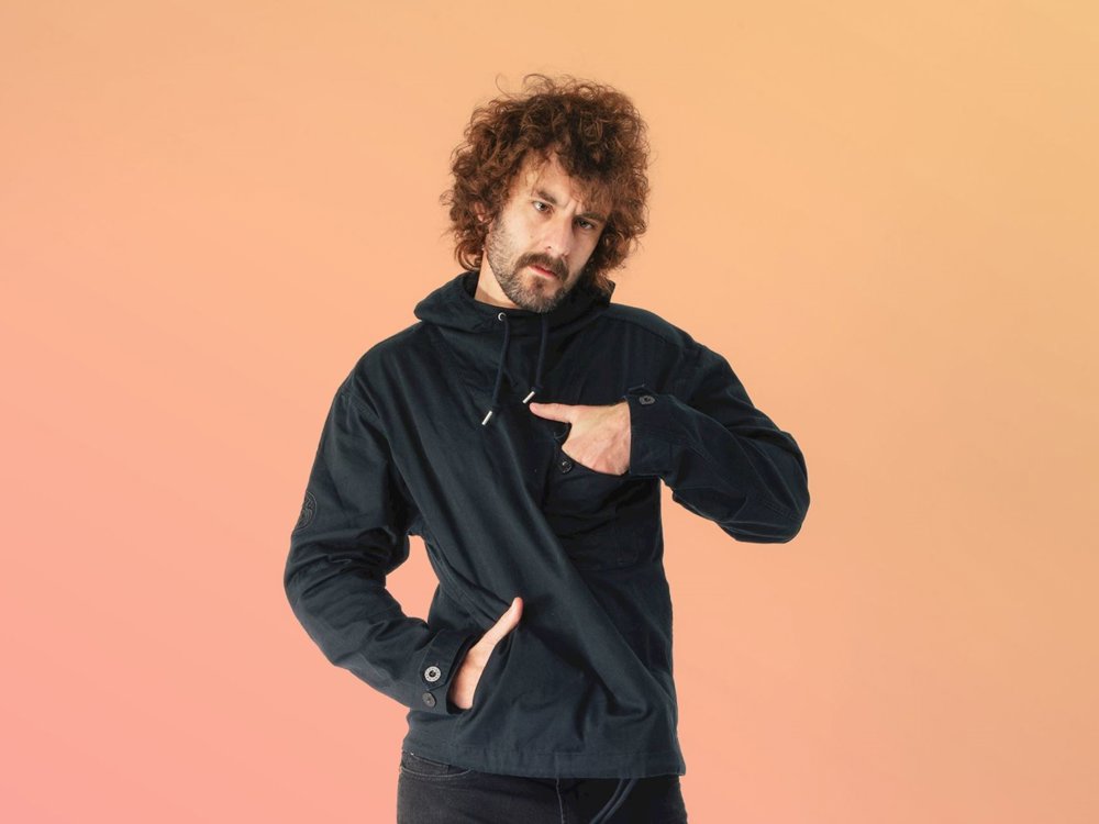 A man with curly hair and a beard points to the chest pocket of his dark hooded jacket while looking directly at the camera. He stands against a soft peach-to-orange gradient background.
