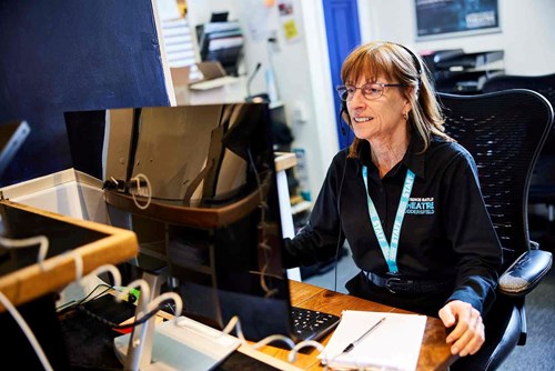 A member of the Lawrence Batley Theatre box office team sits at a desk wearing a headset and staff lanyard, smiling while assisting a customer on the computer.