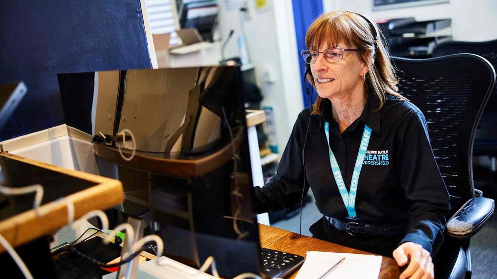 A member of the Lawrence Batley Theatre box office team sits at a desk wearing a headset and staff lanyard, smiling while assisting a customer on the computer.