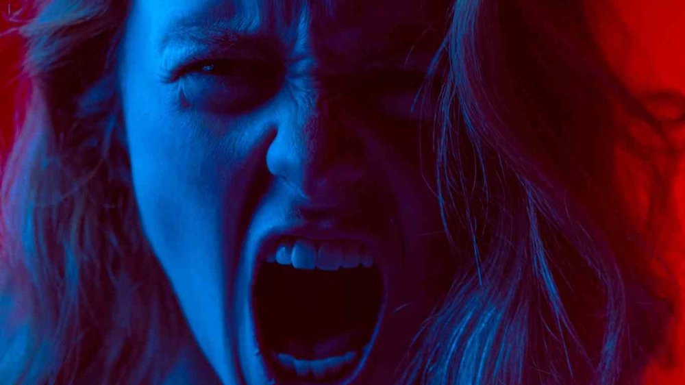 A person with long hair bathed in dramatic red and blue lighting, screaming or shouting with intense emotion, against a vivid red background.