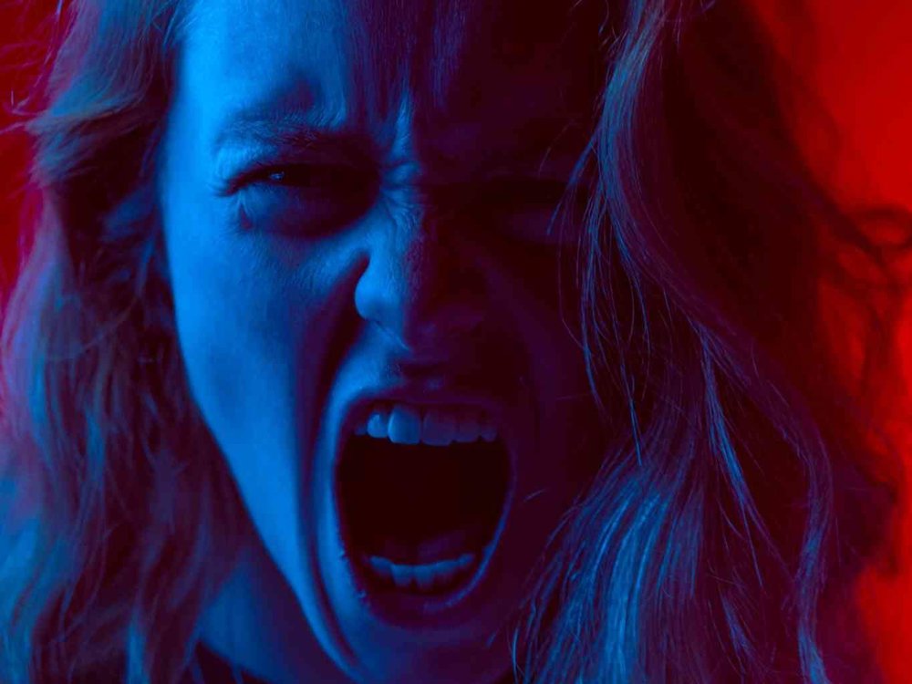 A person with long hair bathed in dramatic red and blue lighting, screaming or shouting with intense emotion, against a vivid red background.