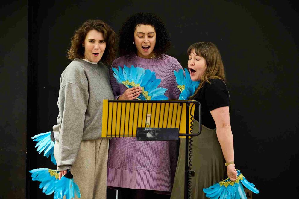 Three actors standing close together during a rehearsal, holding vibrant blue feather fans. They appear to be singing, with a black music stand in front of them. 