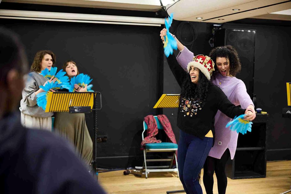 Two actors in the foreground performing energetically, one wearing a crown and black jumper, while holding blue feather fans. In the background, two others stand at music stands with blue fans, engaged in the scene. A rehearsal space with chairs and props is visible. 