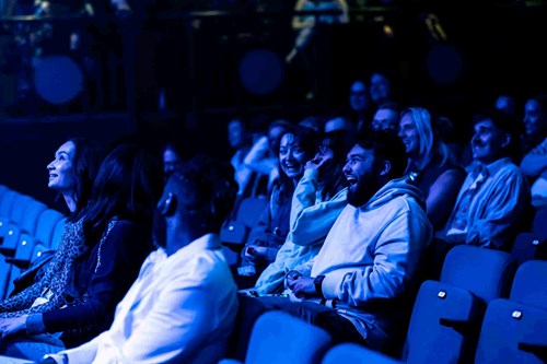 Audience members at Lawrence Batley Theatre laugh and smile as they watch a performance, illuminated by atmospheric blue stage lighting.