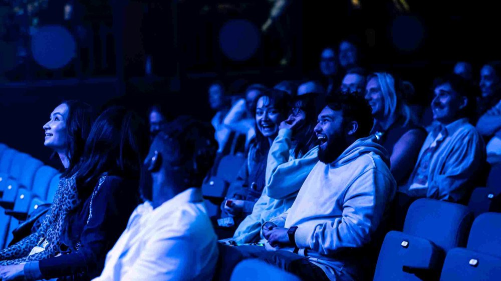 Audience members at Lawrence Batley Theatre laugh and smile as they watch a performance, illuminated by atmospheric blue stage lighting.