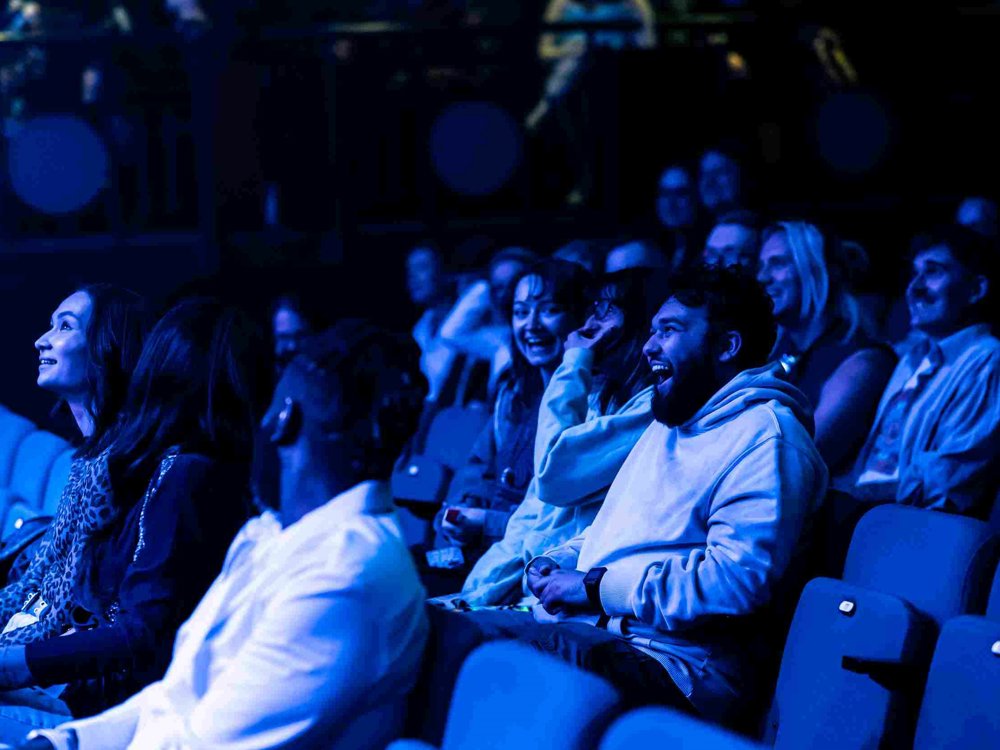 Audience members at Lawrence Batley Theatre laugh and smile as they watch a performance, illuminated by atmospheric blue stage lighting.