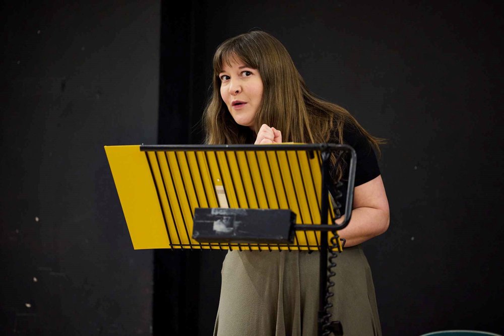 An actor standing behind a black music stand holding a yellow folder, delivering lines with an animated expression. 