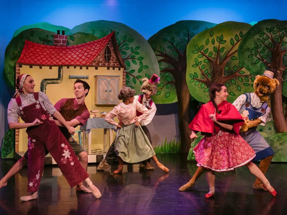 The cast of dancers perform in front of a painted woodland and cottage backdrop, including Little Red Riding Hood, the Wolf, Grandma, and other characters mid-dance.