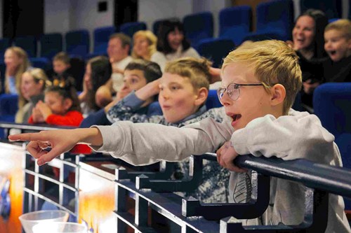 A boy wearing glasses excitedly points towards the stage from a theatre balcony, his face lit with amazement. Around him, other children and adults watch the show with joyful expressions.