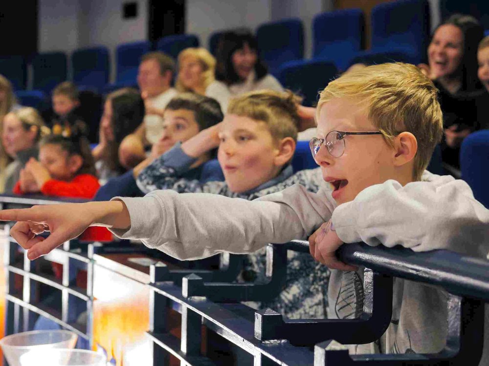 A boy wearing glasses excitedly points towards the stage from a theatre balcony, his face lit with amazement. Around him, other children and adults watch the show with joyful expressions.