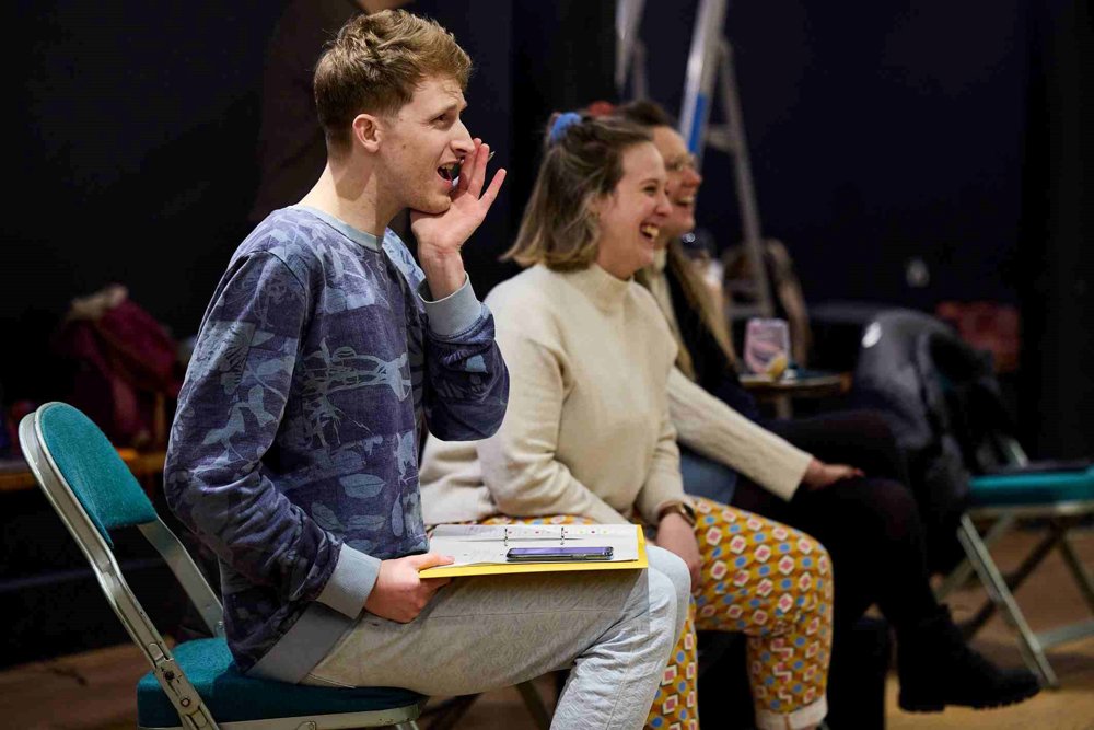 A lively rehearsal scene with three people seated. One person in a patterned sweatshirt is cupping their hand to their mouth, another in a cream jumper and patterned trousers is laughing, and a third person is slightly obscured in the background.
