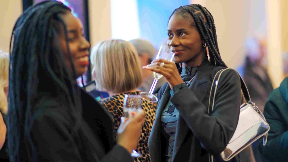 Guests enjoy drinks in the foyer at Lawrence Batley Theatre. One woman smiles while holding a glass, and another sips from a champagne flute, surrounded by a lively crowd.