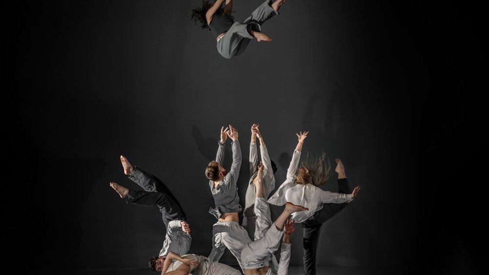 A dynamic performance by a group of contemporary dancers from Motionhouse, showcasing acrobatic choreography. One dancer is mid-air in a dramatic flip, while others form intricate, intertwined shapes on the ground, reaching upward.