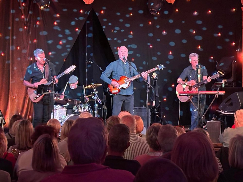 Four-piece band performing on stage with guitars and drums in front of a seated audience, with warm lighting and dotted light effects behind them.