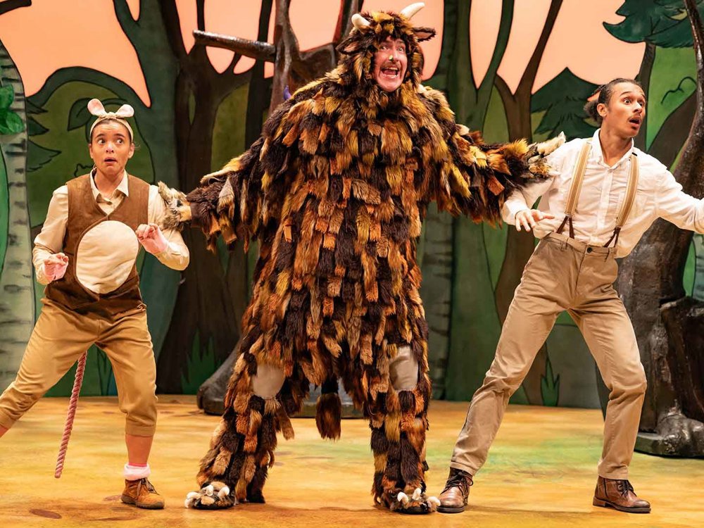Three actors on a forest-themed stage. A large, shaggy creature with horns stands in the center with arms outstretched, while two smaller animal characters on either side react with surprise.