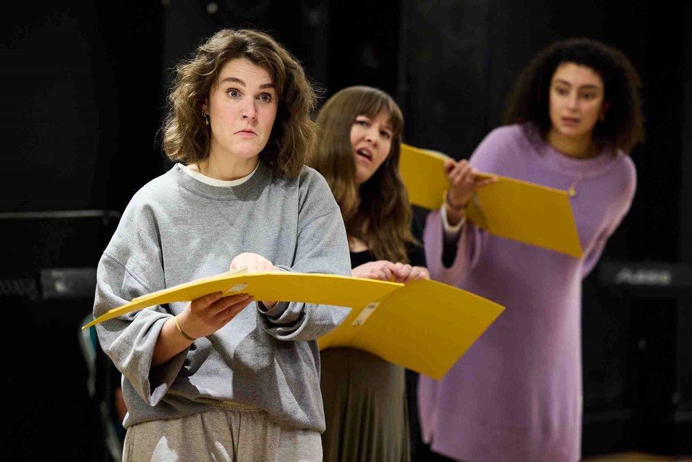 Three actors in a rehearsal space holding yellow folders, standing in a line. The actor in the foreground wears a grey sweatshirt and looks intently, while the two behind follow along, with one wearing a purple jumper and another in a black top.