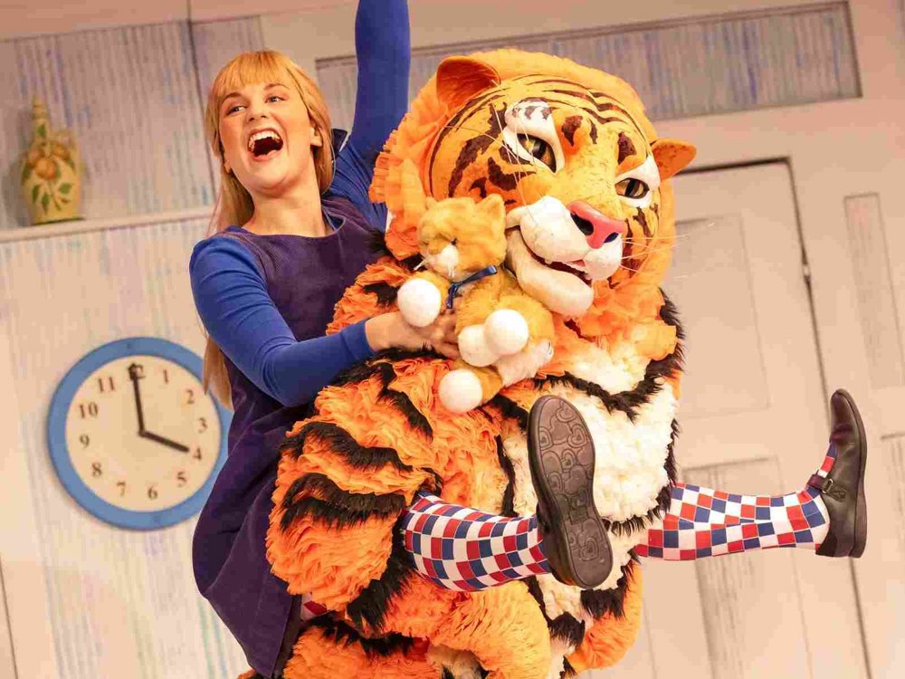 A joyful young woman rides on the back of a large tiger character, raising one arm in celebration while holding a teddy bear, on a bright theatrical kitchen set.