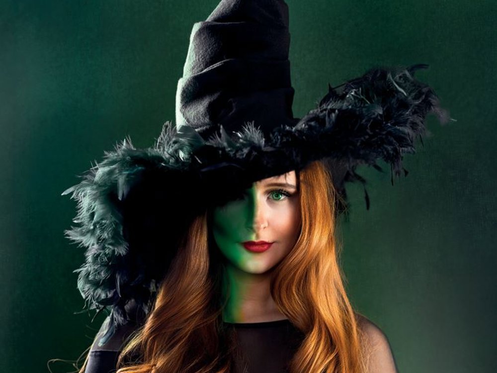 A young woman with long red hair wears a dramatic black witch hat trimmed with feathers, partially shadowing her face against a dark green background.