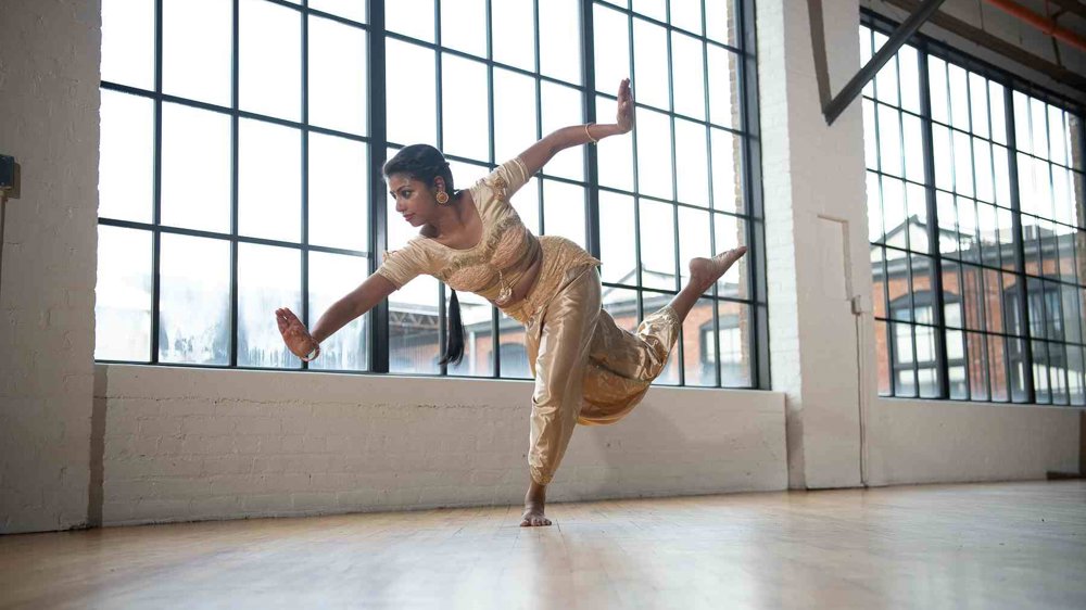 A dancer in a gold traditional Indian costume balances gracefully on one foot, arms extended and one leg raised behind her, in a bright industrial-style studio with large grid windows and wooden floors.