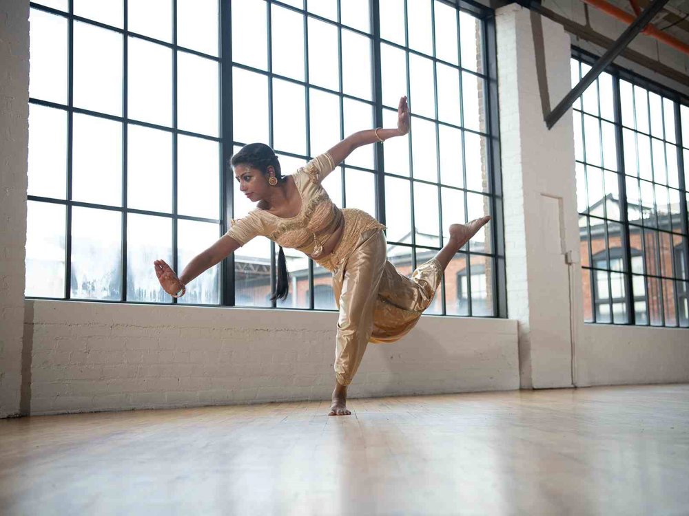 A dancer in a gold traditional Indian costume balances gracefully on one foot, arms extended and one leg raised behind her, in a bright industrial-style studio with large grid windows and wooden floors.