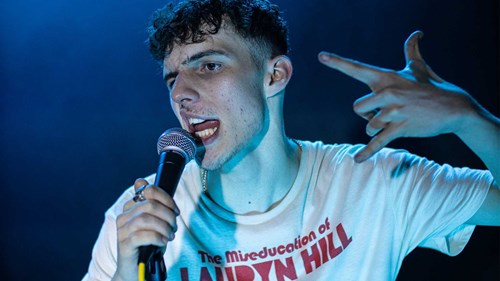 A young man is speaking into a microphone with intensity, wearing a white T-shirt that reads 'The Miseducation of Lauryn Hill.'