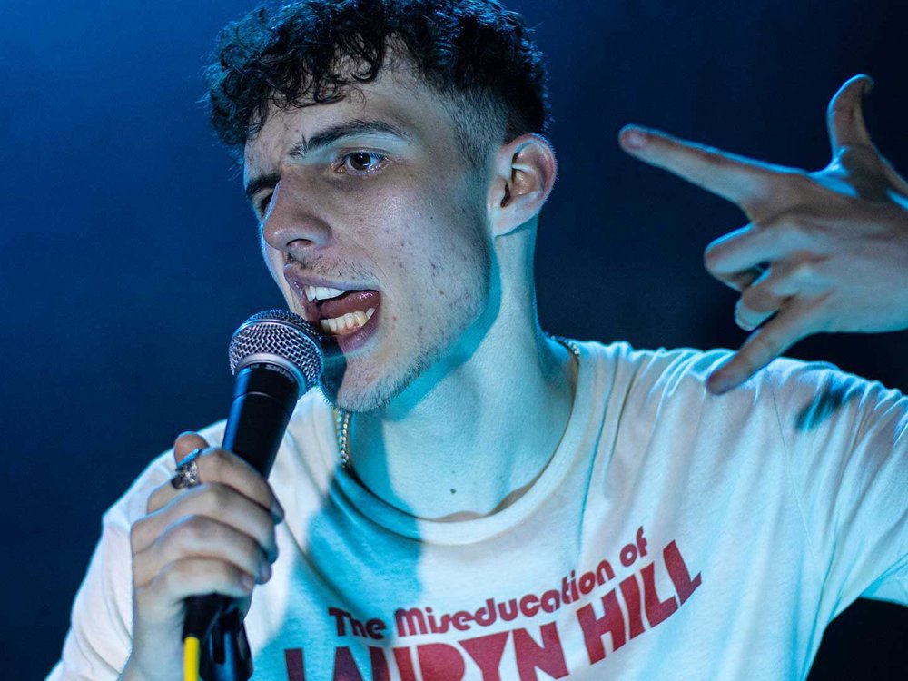 A young man is speaking into a microphone with intensity, wearing a white T-shirt that reads 'The Miseducation of Lauryn Hill.'