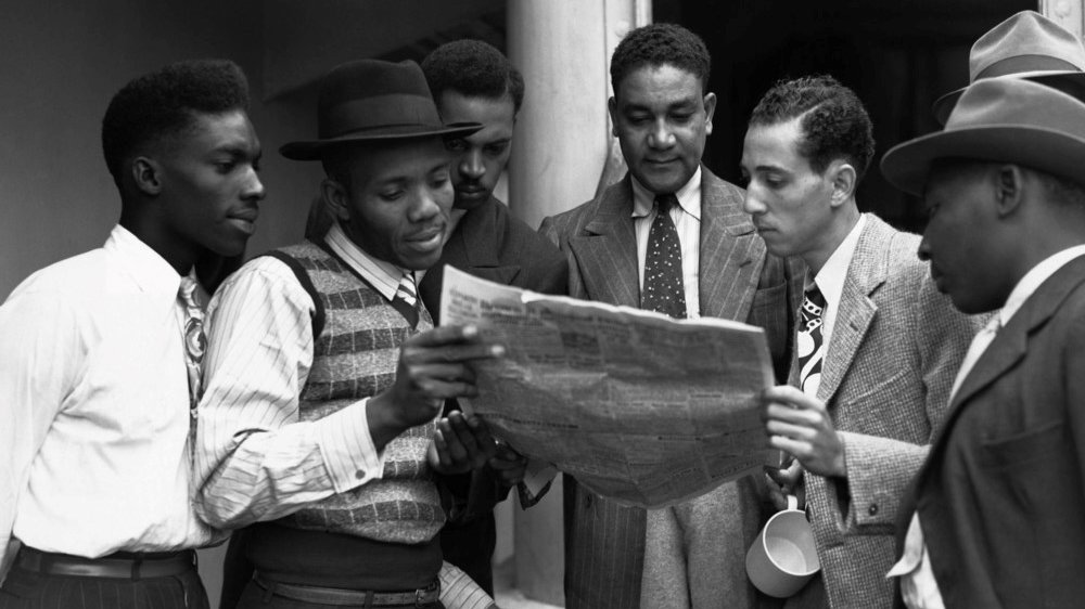 Black-and-white photograph of six young men gathered outside a building, closely reading a newspaper together.