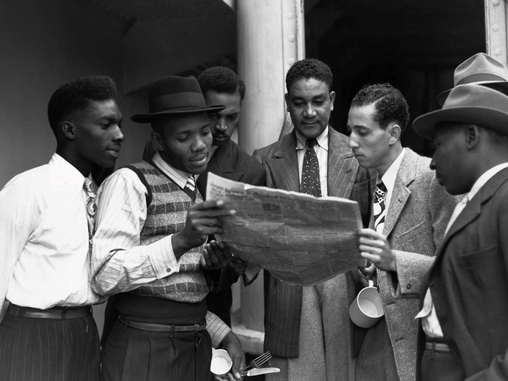Black-and-white photograph of six young men gathered outside a building, closely reading a newspaper together.