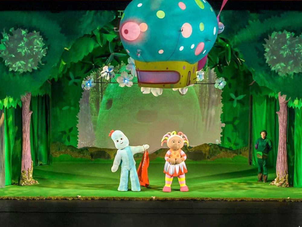 Igglepiggle and Upsy Daisy stand centre stage beneath the floating Pinky Ponk, with audience members visible in the foreground.
