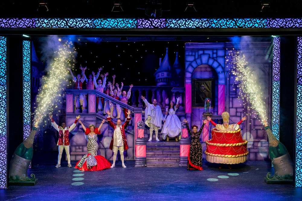 Pantomime cast celebrate on a grand fairytale stage with arms raised as sparkling pyrotechnic effects erupt on either side, framed by a castle set, starry backdrop, and colourful theatrical lighting.