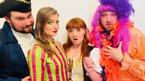 Four panto actors in colourful costumes pose dramatically, including a dame in a wig and feather boa.