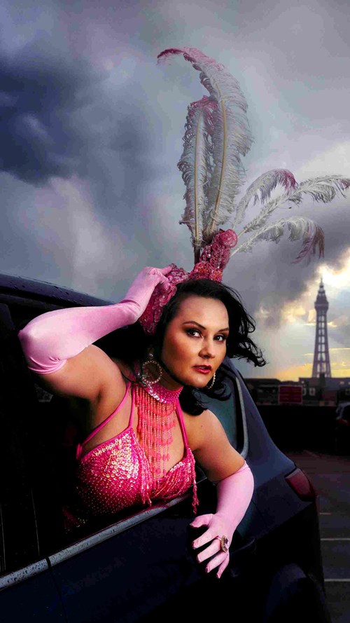 Rachel Fairburn poses out of a car window, dressed in a bright pink dress with pink gloves and a headdress. 