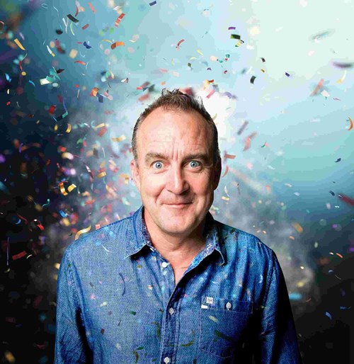 A man with short, slightly spiky brown hair and bright blue eyes is smiling with a playful expression. He is wearing a blue button-up shirt with a subtle pattern and a small lapel pin. The background is a dynamic, vibrant mix of blue and white light with multicoloured confetti falling around him, creating a celebratory atmosphere