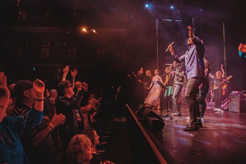 A lively stage performance with singers and musicians engaging an enthusiastic audience. A lead vocalist stands at the front with a microphone, arms raised, while the audience claps and cheers along.