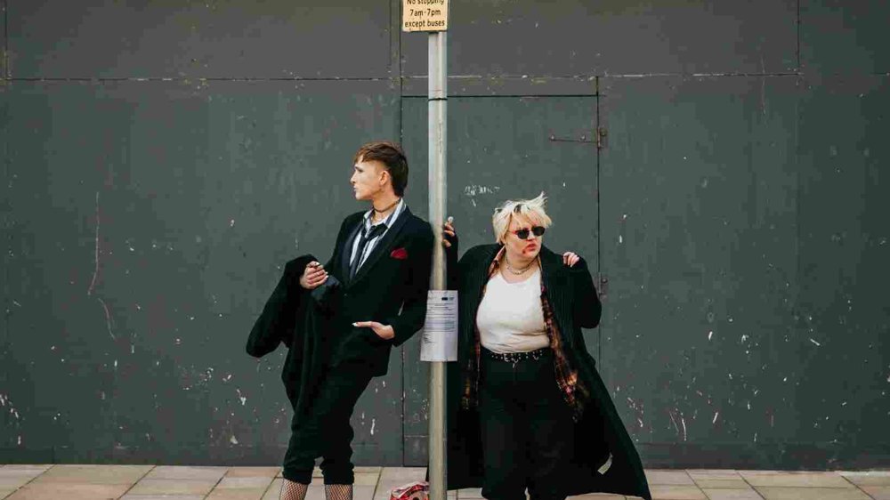 Two people stand at a bus stop on a city pavement, dressed in bold, alternative clothing. One leans against the bus stop pole in a suit and boots, while the other wears sunglasses, a long coat, and checked shirt. A backpack sits on the ground between them against a backdrop of an urban wall.