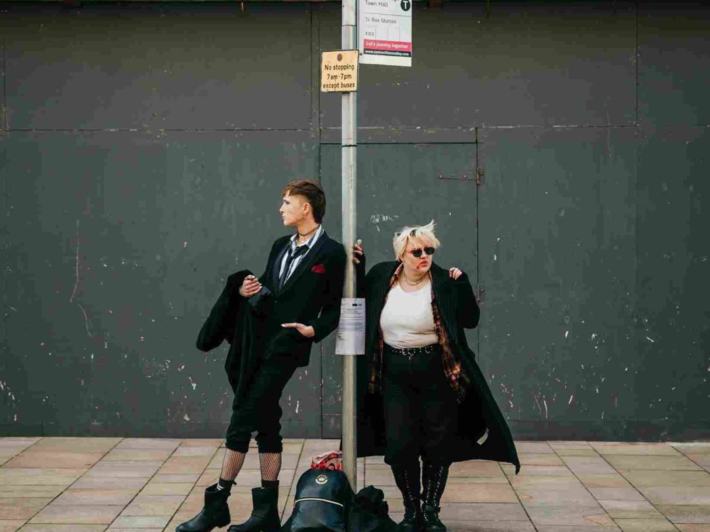 Two people stand at a bus stop on a city pavement, dressed in bold, alternative clothing. One leans against the bus stop pole in a suit and boots, while the other wears sunglasses, a long coat, and checked shirt. A backpack sits on the ground between them against a backdrop of an urban wall.