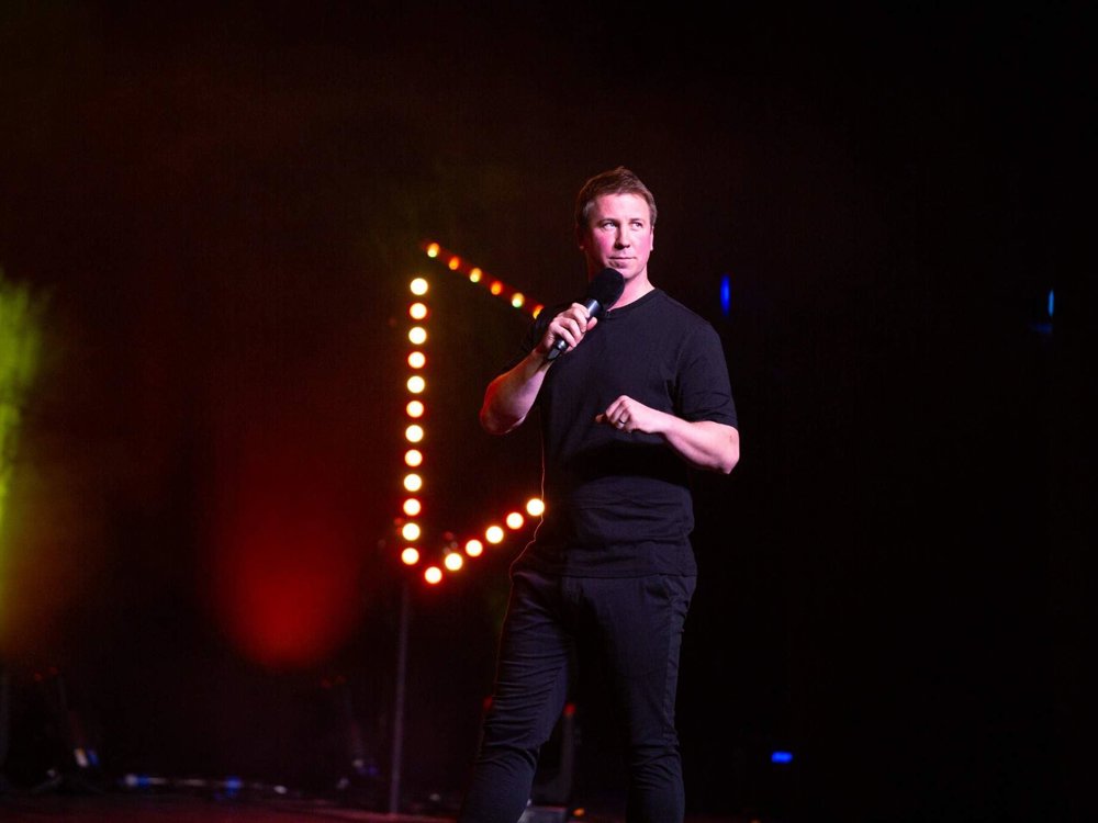 A male stand-up comedian in a black outfit holding a microphone on stage, lit by dramatic red and yellow stage lights with a triangular light rig in the background