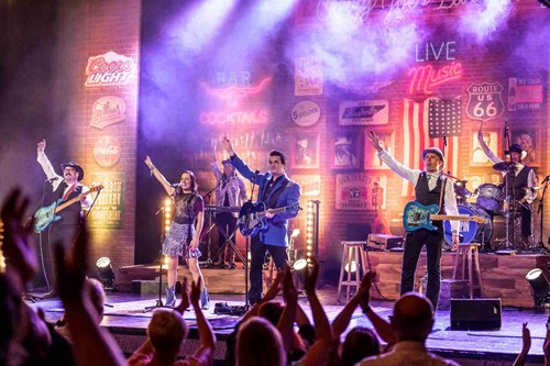 A full band performs on stage in front of a vibrant, Americana-themed backdrop with neon signs and a large U.S. flag. All performers raise their arms in unison as the audience claps along in the foreground.