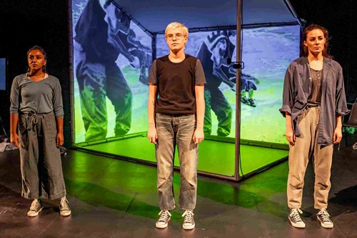 Three young people stand, spaced slightly apart, looking ahead of them. Behind them there is a projection.