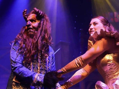 Belle and the Beast, from Beauty and the Beast, hold hands on stage. The Beast is wearing special effects makeup, with horns and a beast like face.