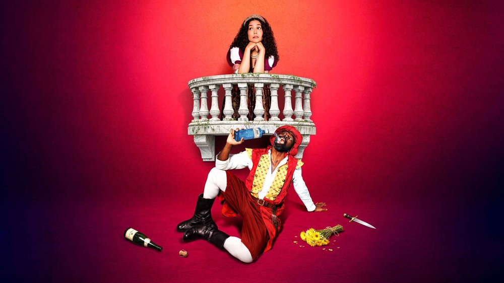 A theatrical promotional image showing a woman leaning over a stone balcony while a man in a red Renaissance-style costume sits below drinking from a bottle. A bouquet of flowers, a discarded bottle and a dagger lie on the floor against a bright pink background.