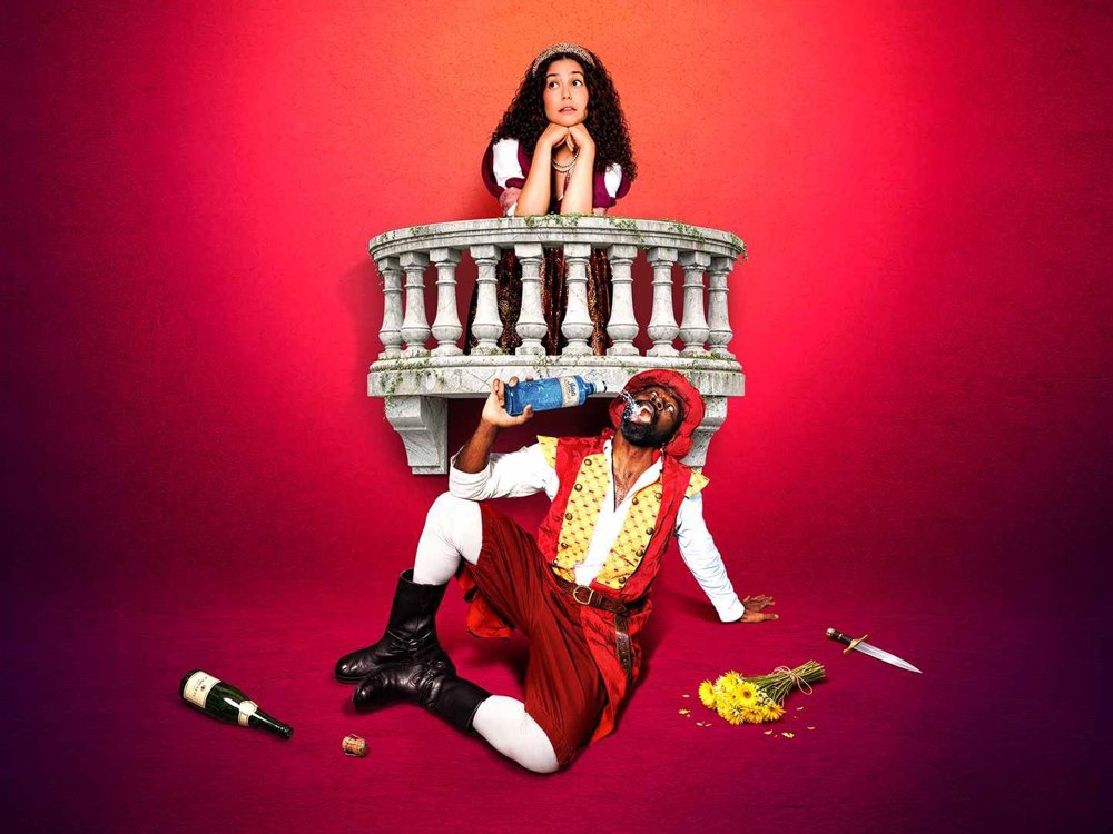 A theatrical promotional image showing a woman leaning over a stone balcony while a man in a red Renaissance-style costume sits below drinking from a bottle. A bouquet of flowers, a discarded bottle and a dagger lie on the floor against a bright pink background.