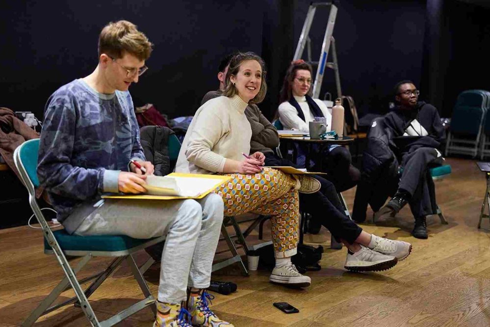 A group of five people seated in a semi-circle during a rehearsal. Two are holding scripts, one in a patterned sweatshirt and another in a cream jumper with colourful trousers, while others are watching attentively in the background.