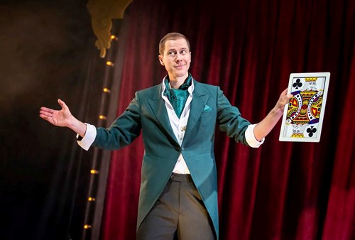 A character dressed smartly in a green suit is holding an oversized playing card of the King. 