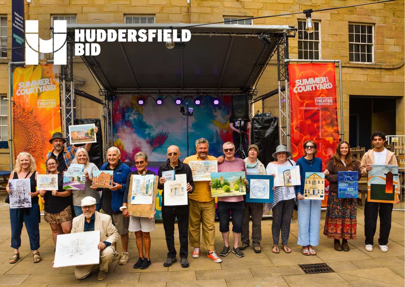 Group of artists stand in front of an outdoor stage branded Huddersfield BID, each holding an original artwork, with Summer Courtyard banners and historic stone buildings behind them.
