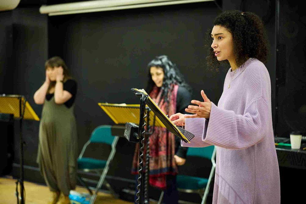 Three actors during a rehearsal, each standing at music stands with scripts. The actor in the foreground, wearing a lavender jumper, gestures passionately while speaking, while the others react behind her. 