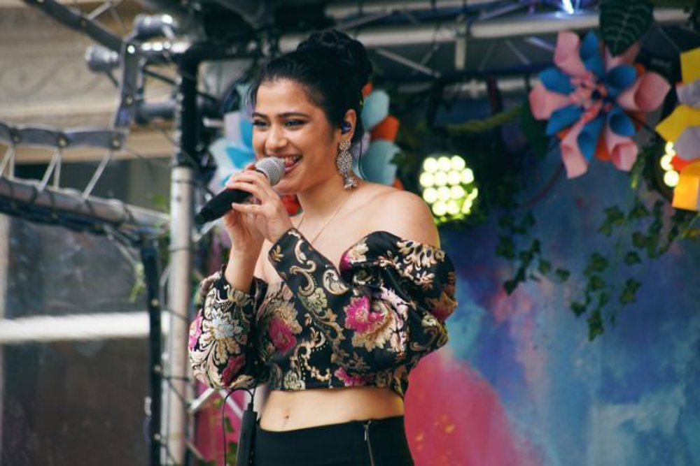 A smiling singer holding a microphone on an outdoor stage, dressed in an elegant floral-patterned off-shoulder top. Bright stage lights and colourful decorations provide a festive backdrop.